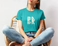 A woman sitting in a chair wearing an Aloha Maluhia In Teal t-shirt featuring the Be Still and Know logo, representing Christian apparel.