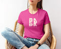 A woman wearing a Golden Coast Vibes Tee In Berry, adorned with the Be Still and Know Logo, sitting in a chair.