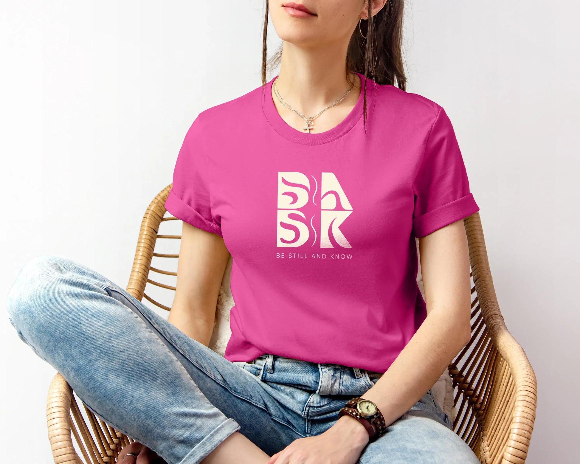 A woman wearing a Golden Coast Vibes Tee In Berry, adorned with the Be Still and Know Logo, sitting in a chair.