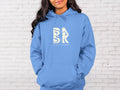 A woman wearing a Coastal Calm Hoodie In Carolina Blue with the Be Still and Know logo on it.