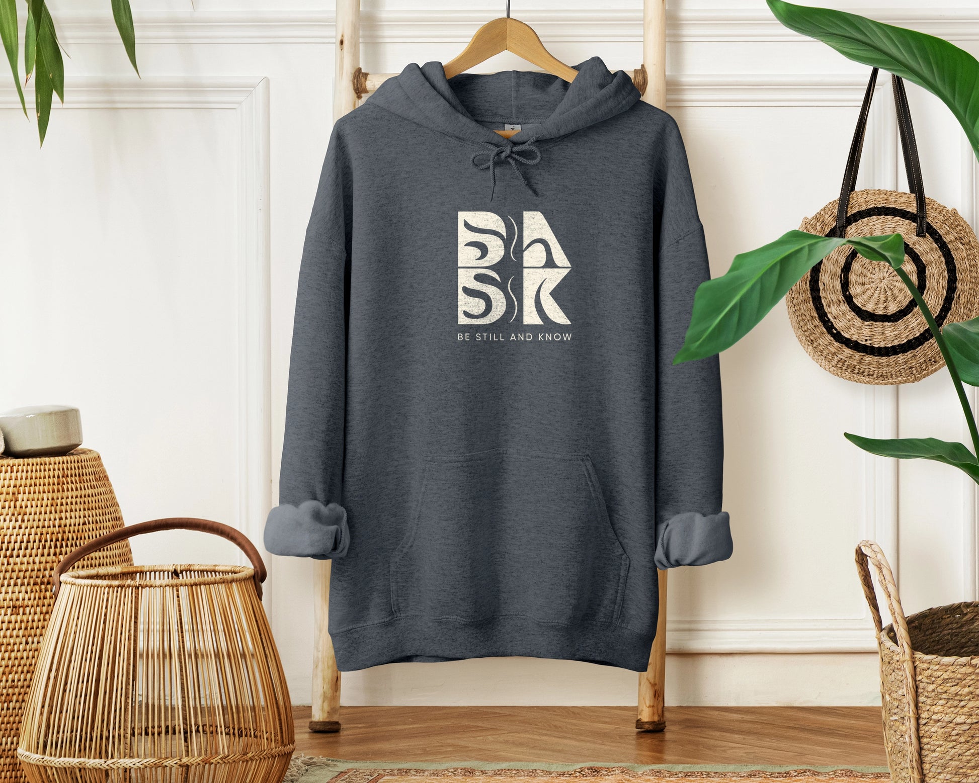 A "Blessed Beginnings Hoodie in Dark Heather" with the word "sark" on it, featuring the "Be Still and Know" logo.