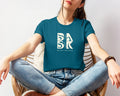 A woman sitting in a chair wearing a teal t-shirt with the Be Still and Know logo on it, called Blessed Beginnings In Deep Teal.