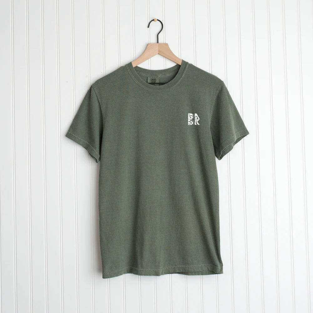 Olive green God Means It For Good Shirt with a small logo symbolizing "God grows new life" on the chest, hanging on a wooden hanger against a white vertical panel background by Be Still and Know.