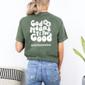 A woman stands with her back turned, displaying a green God Means It For Good Shirt from Be Still and Know, with a floral design and additional text underneath.