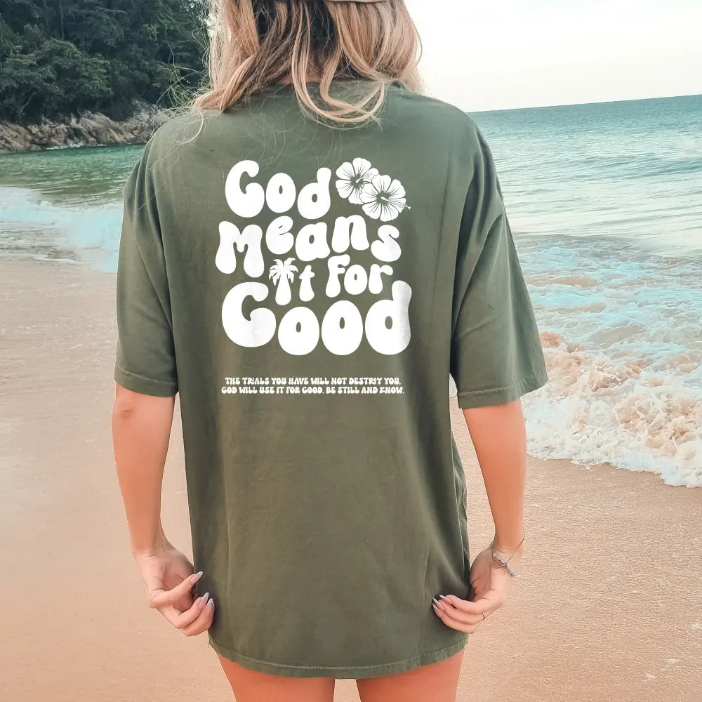 Person standing on a beach wearing a Be Still and Know God Means It For Good Shirt, colored green with the text "God means it for good" along with a floral design and additional text below.