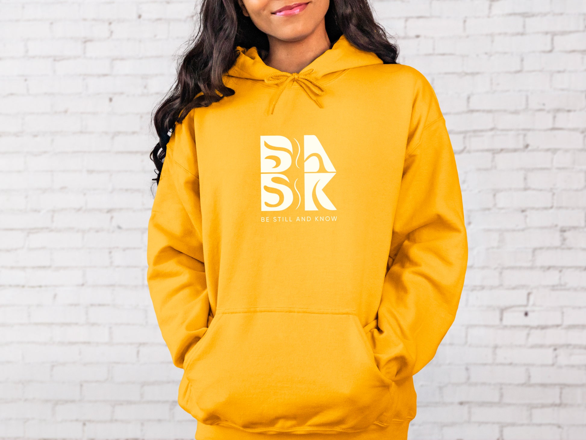 A woman wearing a yellow hoodie with the word Golden Coast In Hoodie In Gold on it, showcasing Be Still and Know logo.