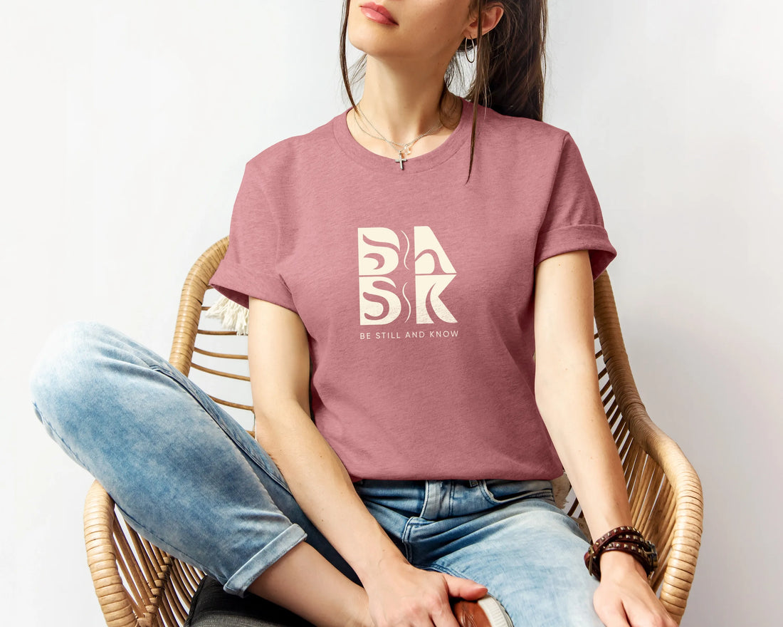 A Coastal Calm Tee In Heather Mauve by Be Still and Know with the word bask on it.