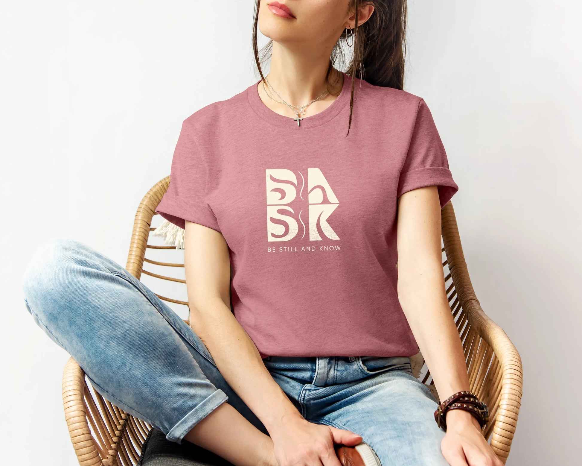 A woman wearing a Coastal Calm Tee In Heather Mauve by Be Still and Know logo sitting on a chair, showcasing Christian Apparel.