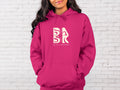 A woman wearing a Golden Coast Hoodie in Heliconia with the Be Still and Know logo on it.