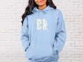 A woman wearing a Coastal Calm Hoodie In Light Blue with the Be Still and Know Logo on it.