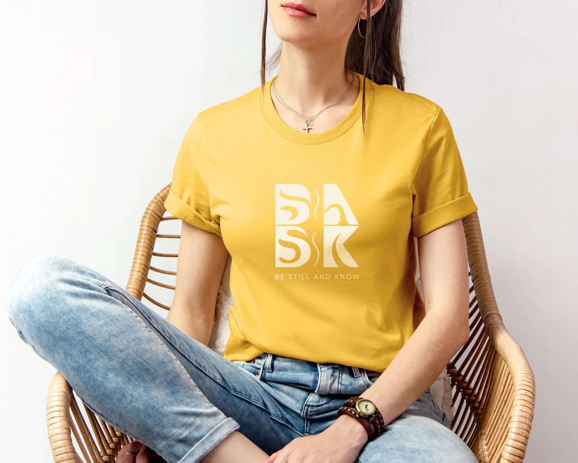 A woman sitting in a chair wearing a yellow t-shirt showcasing the "Golden Coast Tee In Maize Yellow" by Be Still and Know.