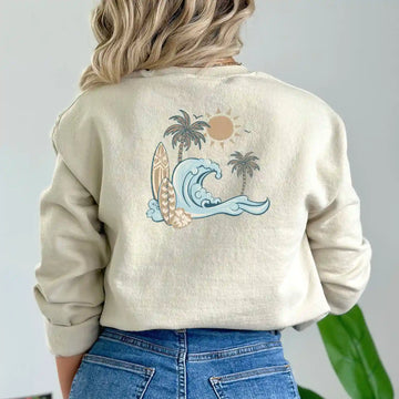 The back of a woman wearing a Nalu o ka Mana (Waves of Faith) sweatshirt with a wave and palm trees, featuring the Be Still and Know logo.