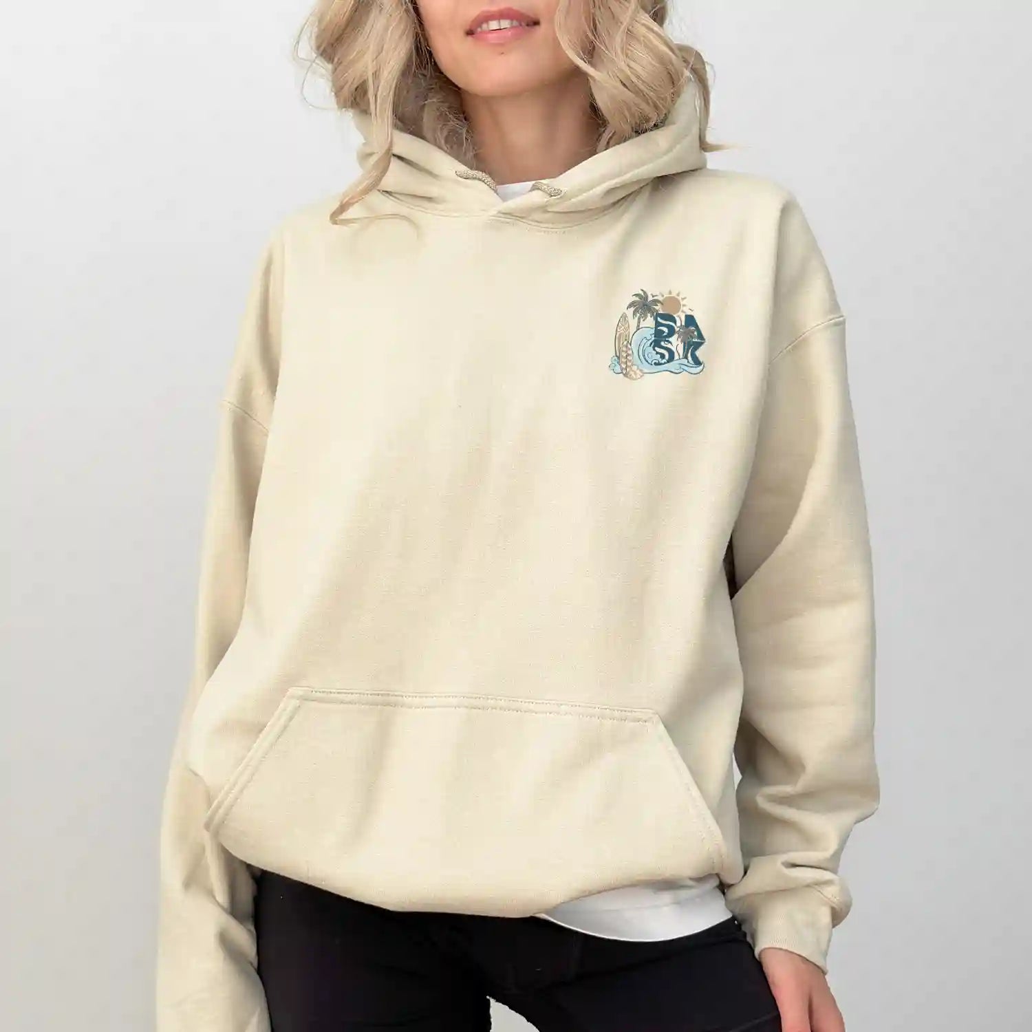 A woman wearing a beige Nalu o ka Mana (Waves of Faith) Hoodie by Be Still and Know, with an embroidered owl on it, showcasing the BSAK Logo.
