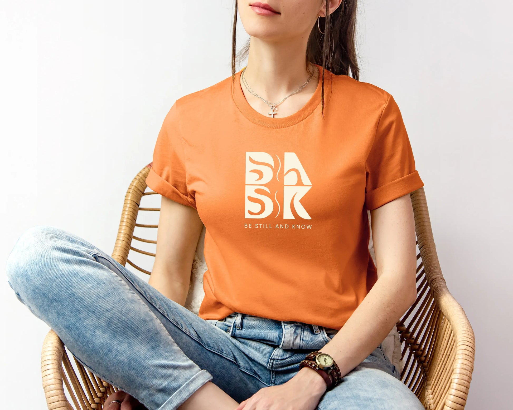 A woman wearing a Golden Coast Tee in Orange featuring the Be Still and Know logo sitting in a chair.