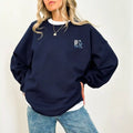 A woman wearing a navy Peace Be Still Sweatshirt and jeans featuring the Be Still and Know Logo.
