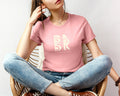 A woman wearing a Coastal Calm Tee In Pink adorned with the Be Still and Know logo, sitting on a chair.