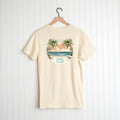 A beige Be Still and Know Salty & Saved shirt with a palm tree and beach graphic, hanging on a wooden hanger against a white wooden background.