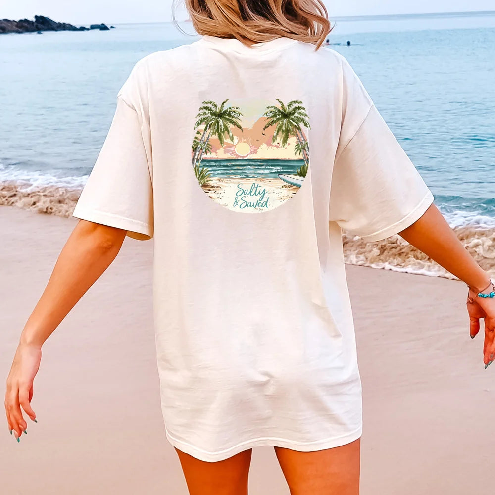 Woman standing on a beach wearing a beige Be Still and Know Salty & Saved Shirt with a circular tropical beach graphic on the back.