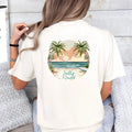 A woman sitting on a couch, seen from behind, wears a white Be Still and Know T-shirt featuring a tropical beach scene graphic with the text 