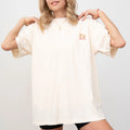 A woman in a cream Be Still and Know Salty & Saved Shirt with a small logo on the chest, touching her collar, against a plain background.