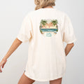 Woman in a white Be Still and Know Salty & Saved Shirt with a tropical beach scene graphic on the back, facing away from the camera, against a plain background.
