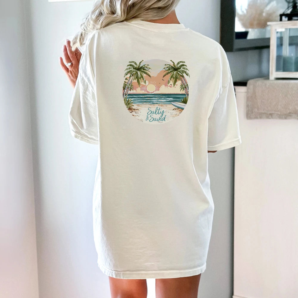 A woman viewed from behind, wearing a white Be Still and Know Salty & Saved Shirt with a colorful tropical beach design printed on the back.