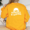 A woman wearing a Sunkissed & Saved Sweatshirt by Be Still and Know that exudes warmth and faith with the phrase 
