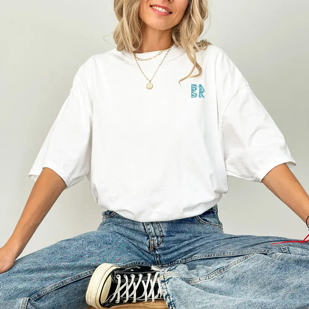 Woman in a Be Still and Know Spirit Lead Me Shirt with a blue monogram and denim jeans, exuding complete trust, sitting down with black sneakers.
