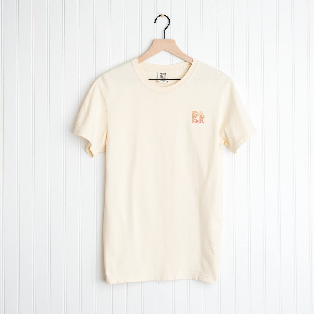 A plain cream Salty & Saved Shirt by Be Still and Know with "br" logo embroidered on the left chest area, hanging on a white hanger against a vertical striped background.