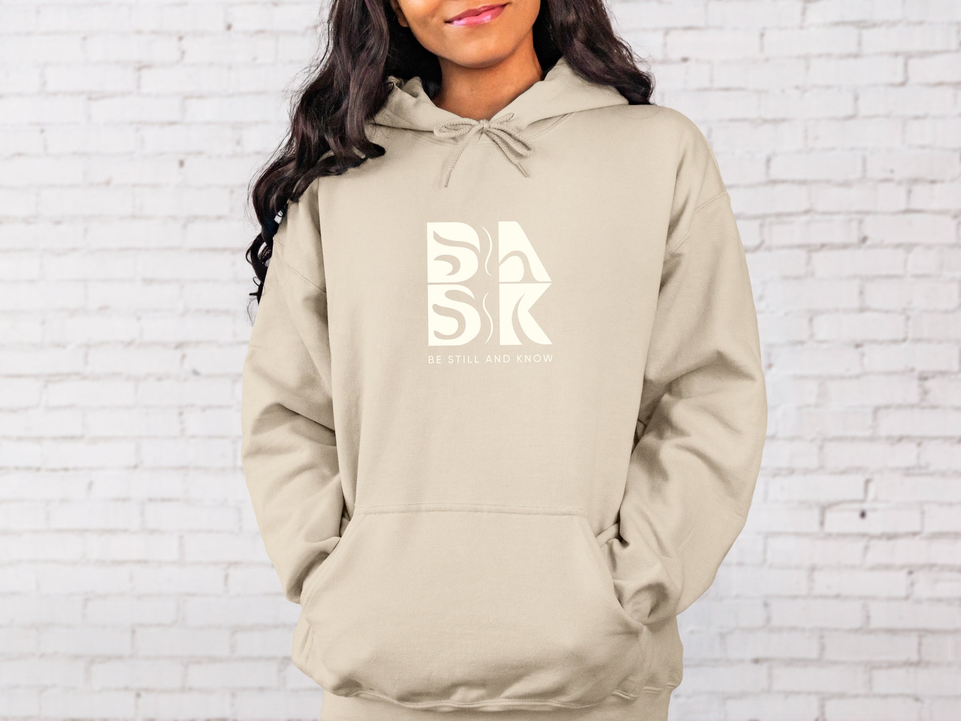 A woman wearing a Coastal Calm Hoodie in Sand with the Be Still and Know logo on it.