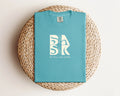 A turquoise Aloha Tee In Seafoam with the Be Still and Know logo prominently featuring the word 