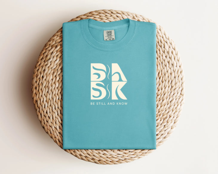 A turquoise Aloha Tee In Seafoam with the Be Still and Know logo prominently featuring the word "bk," perfect for Christian Apparel.