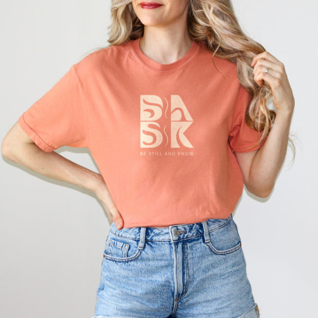 A woman wearing a Aloha Tee In Terracotta by Be Still and Know and denim shorts featuring the BSAK logo.