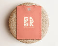 A Be Still and Know Aloha Tee In Terracotta featuring the BSAK logo on a pink background.