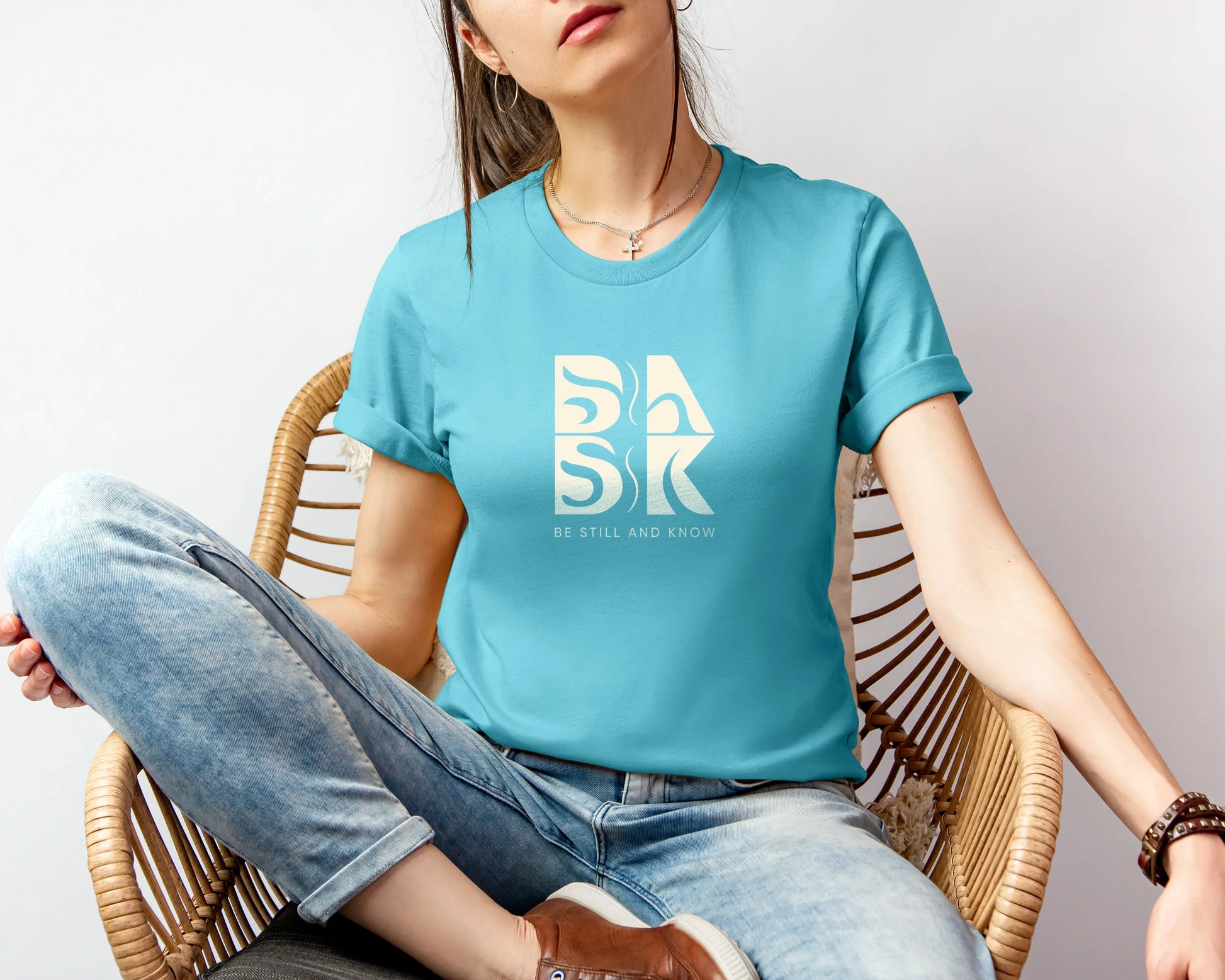 A woman sitting in a chair wearing a blue t-shirt with the Be Still and Know Logo.