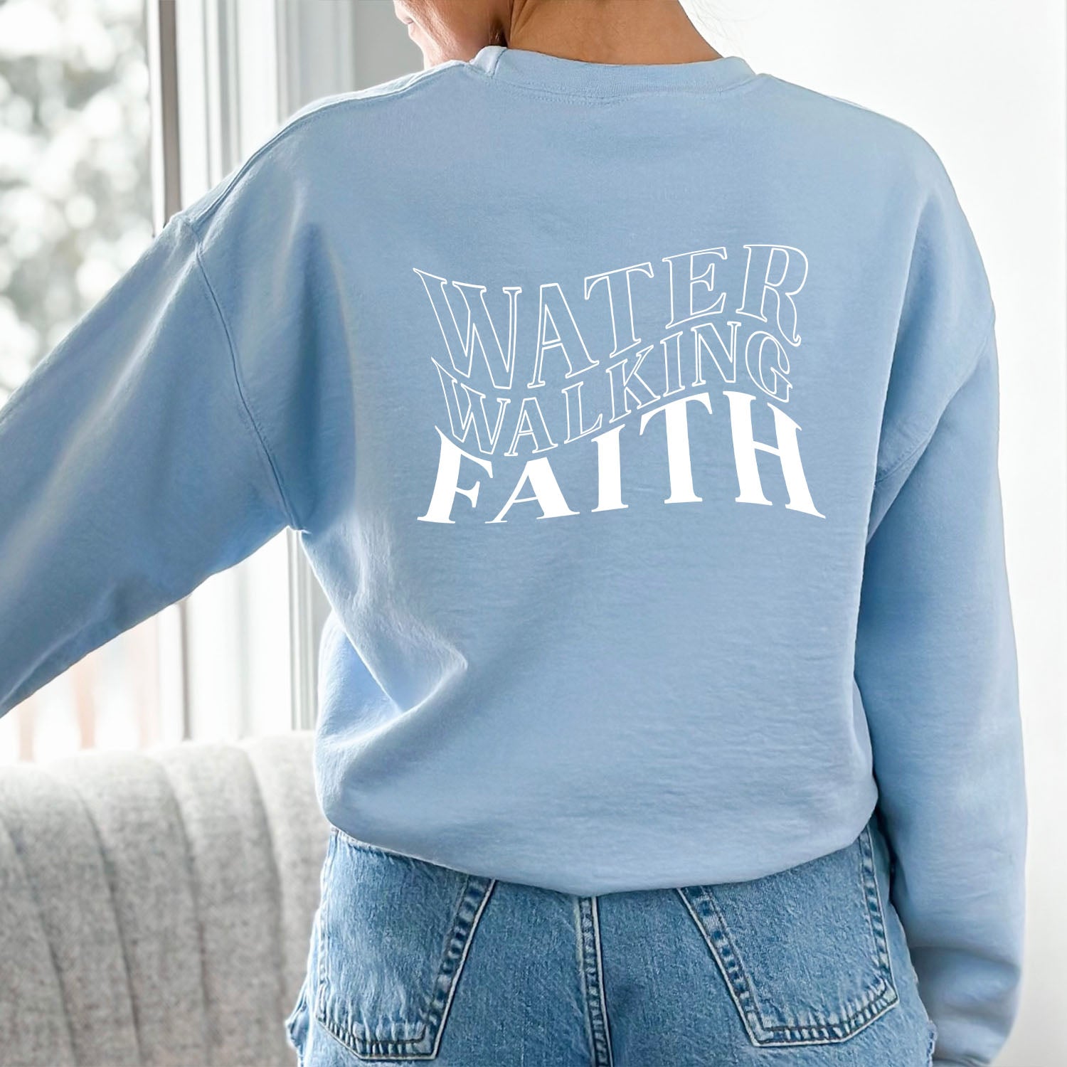 A woman wearing a Water Walking Faith Sweatshirt by Be Still and Know is seen standing in front of a window.