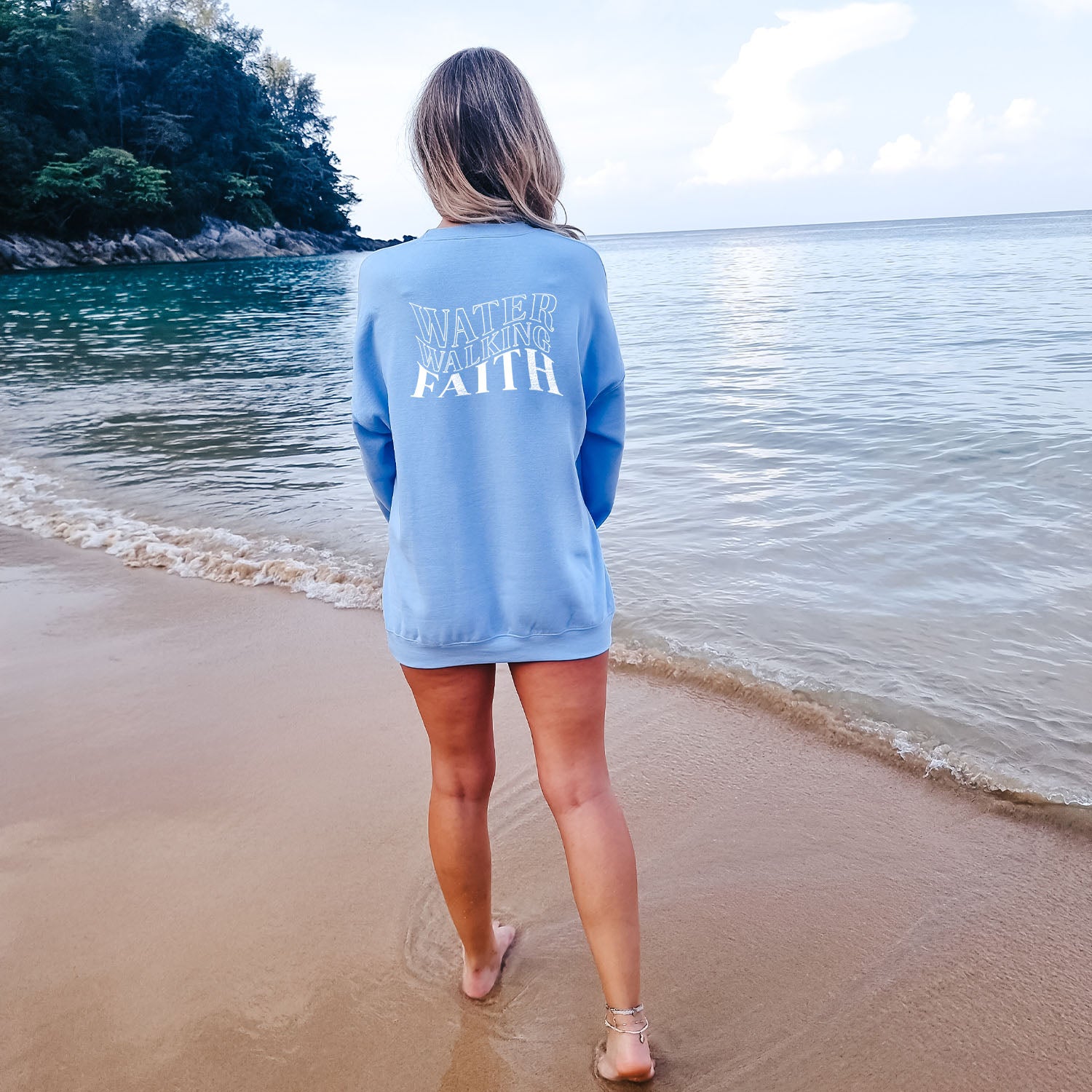 A woman, wearing a Water Walking Faith sweatshirt from the Be Still and Know brand, standing on a beach.