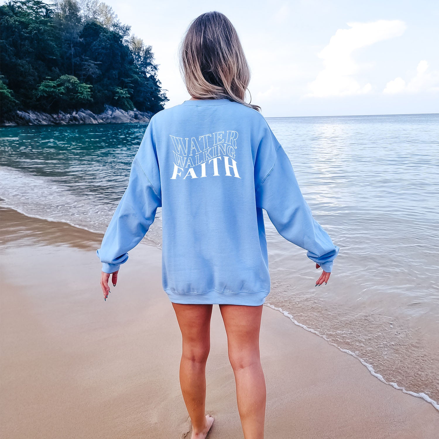A woman, wearing a Water Walking Faith Sweatshirt by Be Still and Know, standing on a beach.