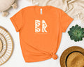 A Golden Coast Tee In Orange with the word bak on it, featuring a Be Still and Know logo.